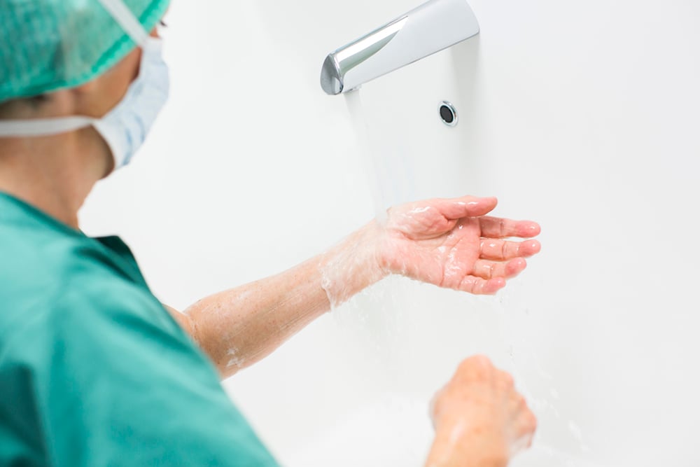 Ansatt kledd i grønt med munnbind vasker hendene.