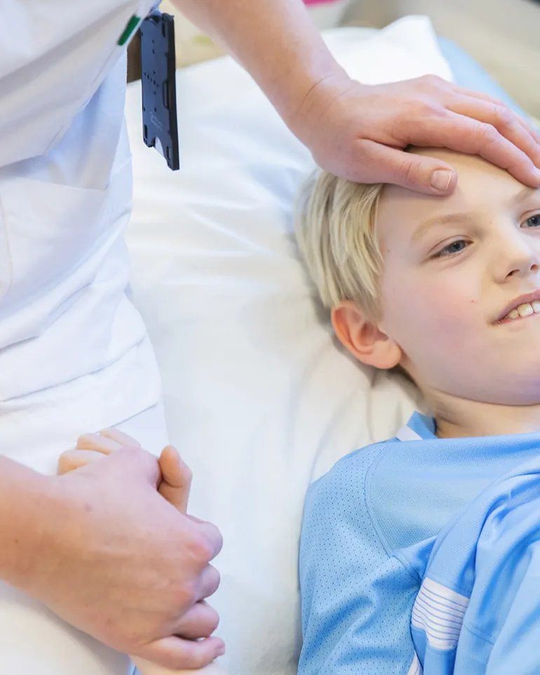 Sykepleier holder et barn i sykehusseng i hånda.