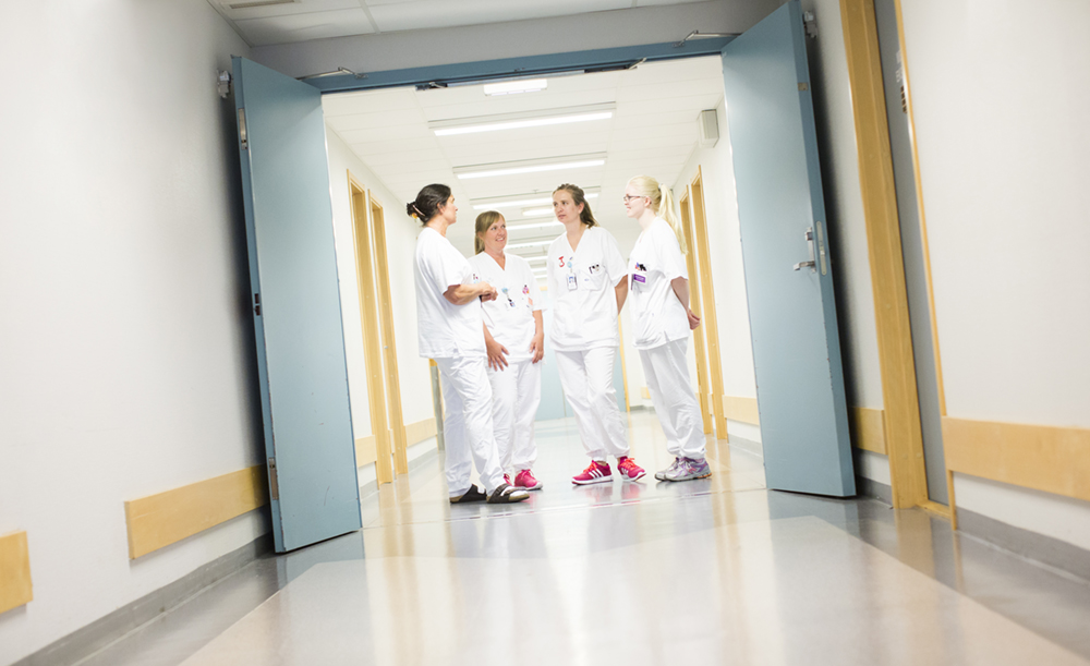En gruppe helsepersonell i hvite uniformer snakker sammen i sykehuskorridor.