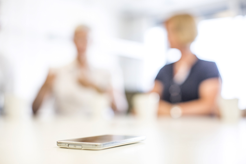 Mobiltelefon ligger på et bord, med to mennesker som snakker sammen i bakgrunnen, illustrasjonsbilde